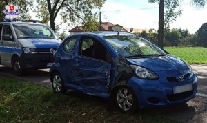 pojazd toyota stojący na poboczu, ma uszkodzony bok, za nim stoj radiowóz policyjny