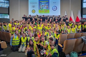 Grupa dzieci w kamizelkach odblaskowych. W tle orkiestra policyjna