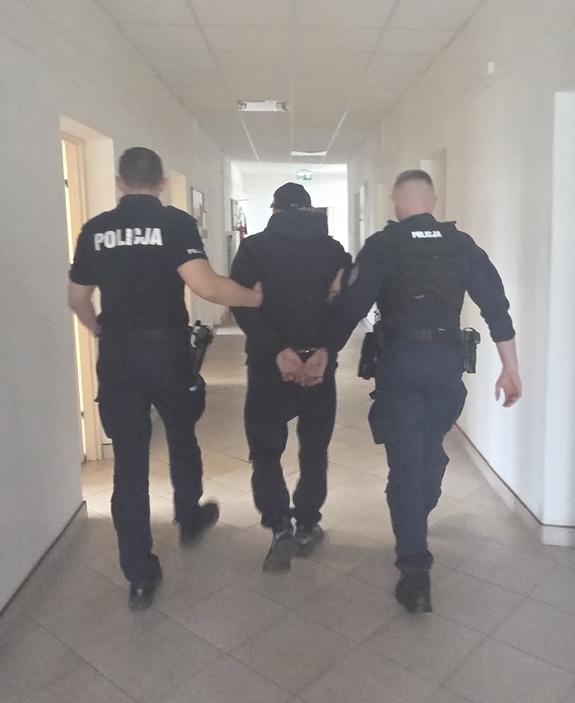 Dwóch policjantów prowadzi przez korytarz mężczyznę, który ma kajdanki założone na ręce trzymane z tyłu